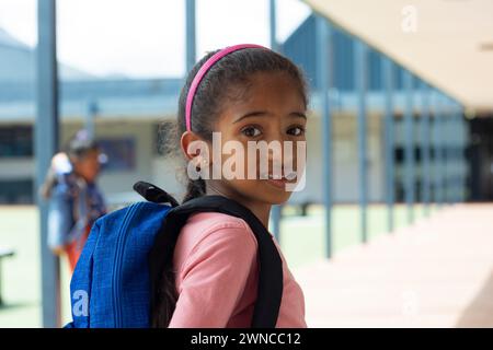 Ein birassisches Mädchen mit rosa Stirnband und blauem Rucksack lächelt in der Schule Stockfoto