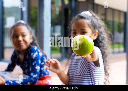 Zwei rassistische Mädchen teilen sich einen Moment in der Schule, eine hält einen grünen Apfel vor die Kamera Stockfoto