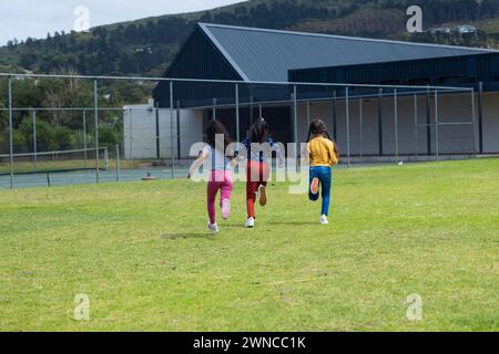 Drei junge Mädchen rennen über ein Grasfeld in der Nähe eines Gebäudes mit Kopierraum in der Schule Stockfoto