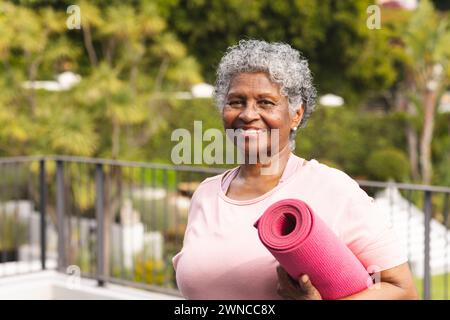 Ältere birassische Frau mit grauen Haaren hält draußen eine rosafarbene Yogamatte Stockfoto