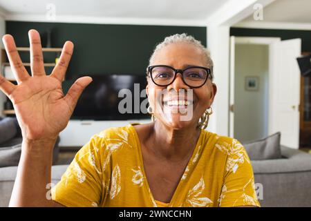 Eine ältere afroamerikanische Frau winkt mit einem hellen Lächeln, trägt eine Brille und ein gelbes Oberteil Stockfoto