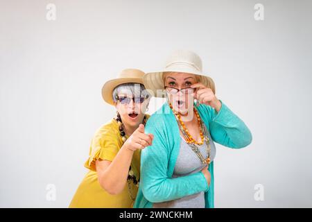 Ältere Freundinnen, die überraschend in die Kamera schauen und mit dem Finger auf grauen Hintergrund zeigen Stockfoto