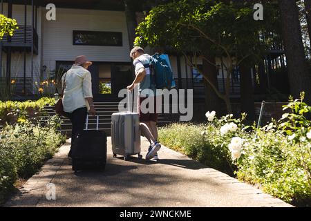 Ein birassisches Paar geht auf ein Haus zu, zieht einen Koffer und trägt einen Rucksack mit Kopierraum Stockfoto