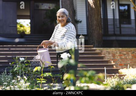 Eine ältere Frau mit grauen Haaren tränkt Pflanzen in einem sonnigen Garten Stockfoto