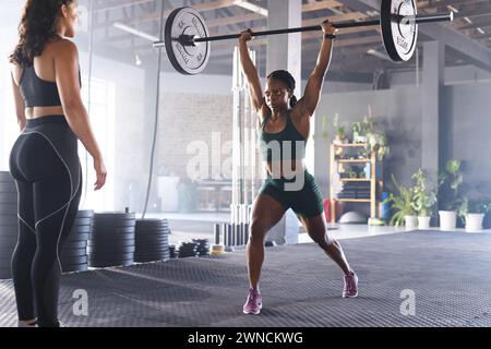 Junge starke birassische Frau hebt Gewichte in einem Fitnessstudio, beobachtet von einer stark fitten birassischen Frau Stockfoto