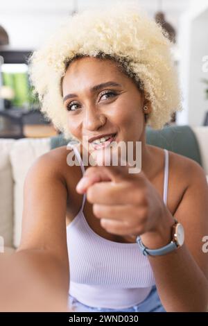 Eine junge birassische Frau mit lockigen blonden Haaren zeigt und lächelt bei einem Videoanruf auf die Kamera Stockfoto