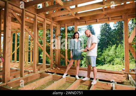 Der Designer zeigt der Frau den Plan, ein Haus aus Holz zu bauen. Eine Investorin inspiziert ihre zukünftige Wohnung in der Nähe des Waldes. Das Konzept der zeitgenössischen ökologischen Konstruktion. Stockfoto