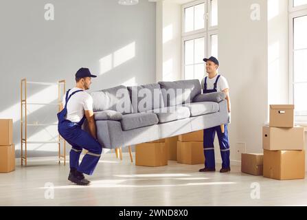 Zwei junge, umziehende Servicemitarbeiter, die ein schweres Sofa zusammen im Wohnzimmer tragen. Stockfoto