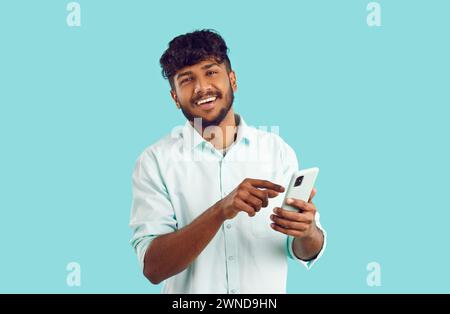 Junger indischer Mann, der Handy hält und lächelnd auf Kameraständer im türkisfarbenen Studio schaut Stockfoto