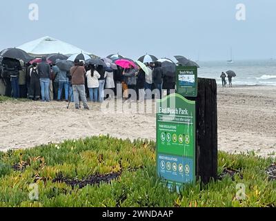 1. März 2024, Santa Barbara, Kalifornien, USA: Während eines weiteren Regensturms, der Santa Barbara County am 1. März 2024 nass, drängten sich fast hundert Menschen unter grauem Himmel und Regenschirmen zusammen, zitterten vor Kälte und Trauer im Regen, bei einer Gedenkfeier im East Beach Park für Dane Angus, ein beliebtes Gemeindemitglied, das diese Woche bei einem Trefferrennen getötet wurde, als er mit seinem Roller fuhr. Sein Arbeitgeber Pure Joy Catering sponserte den Service. Es gibt ein grünes Zeichen im Vordergrund und den Pazifischen Ozean im Hintergrund. (Kreditbild: © Amy Katz/ZUMA Press Wire) NUR REDAKTIONELLE VERWENDUNG! Nicht für C Stockfoto