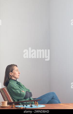 Vertikale Zusammensetzung. Eine zielgerichtete Frau sitzt in einem Stuhl mit überkreuzten Armen über der Brust und blickt aufmerksam nach vorne. In der Nähe auf dem Couchtisch sind Stockfoto