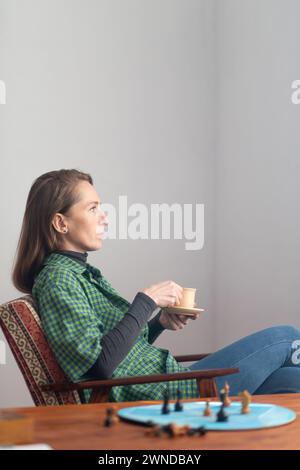 Vertikale Zusammensetzung. Eine Frau sitzt in einem Stuhl mit einer Tasse und Untertasse in der Hand und blickt mit Spannung nach vorne. In der Nähe auf dem Couchtisch befinden sich Schachfiguren Stockfoto