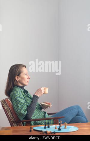 Eine Frau sitzt in einem Stuhl mit einer Tasse und Untertasse in der Hand und blickt mit Spannung nach vorne. In der Nähe auf dem Couchtisch befinden sich Schachfiguren auf einem blauen Kreis. Psy Stockfoto
