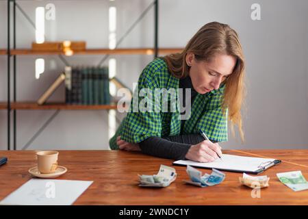 Eine junge hübsche Dame macht nachdenklich und aufmerksam Notizen vor dem Hintergrund von gestaltetem und gezähltem Geld. Stockfoto