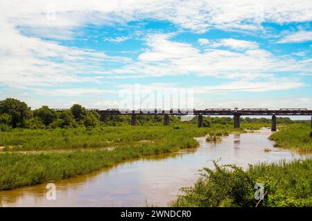 Ein ruhiger Fluss schlängelt sich durch üppiges Grün unter einem riesigen blauen Himmel mit Wolken im Krüger-Nationalpark. Eine alte Eisenbahnbrücke überspannt den W Stockfoto