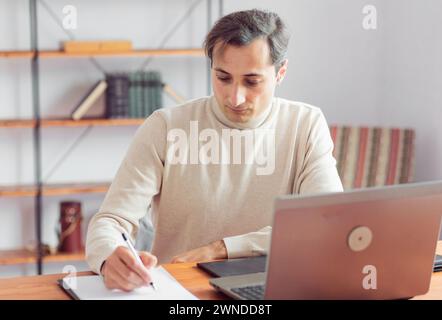 Ein Grafikdesigner im Büro erstellt eine Skizze mit einem Bleistift auf Papier und arbeitet an einem Laptop und einem Grafiktablett. Der Fokus liegt auf dem konzentrierten Gesicht, e Stockfoto