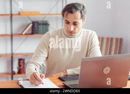 Ein Grafikdesigner im Büro erstellt eine Skizze mit einem Bleistift auf Papier und arbeitet an einem Laptop und einem Grafiktablett. Konzentrieren Sie sich auf ein konzentriertes Gesicht. Stockfoto