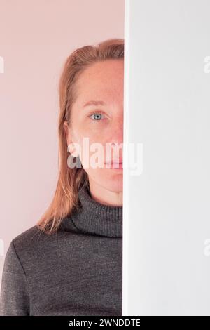 Porträt einer erwachsenen blonden Frau mit grauen Augen. Die Hälfte des Gesichts ist durch eine weiße Wand verdeckt. Stockfoto