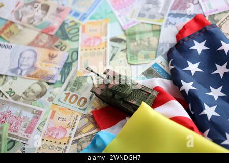 Spielzeugtank auf US-Flagge und ukrainische Flagge auf vielen Banknoten verschiedener Währungen. Hintergrund der Kriegsfinanzierung und militärischen Unterstützung zwischen den Vereinigten Staaten und der Ukraine Stockfoto