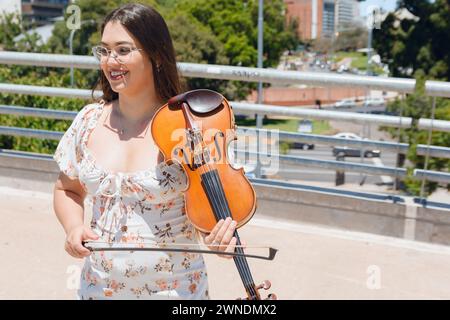 Porträt einer jungen Latina-Buskergeigerin, die mittags auf der Straße stand, unterhielt sich glücklich lächelnd, während sie mit ihrer Geige arbeitete. Stockfoto