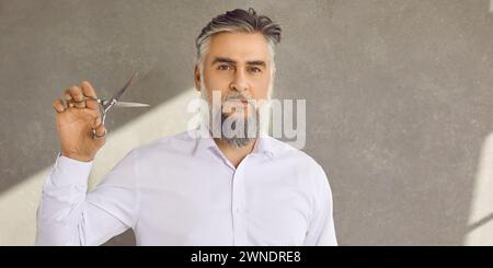 Porträt des bärtigen und mostfarbenen älteren Mannes Friseur mit Schere auf grauem Banner Hintergrund Stockfoto