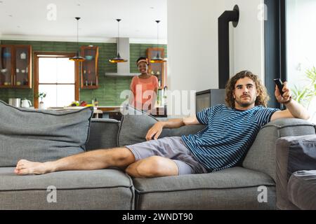 Verschiedene junge Kaukasier sitzen auf einem grauen Sofa, halten eine Fernbedienung, mit lockigen braunen Haaren Stockfoto