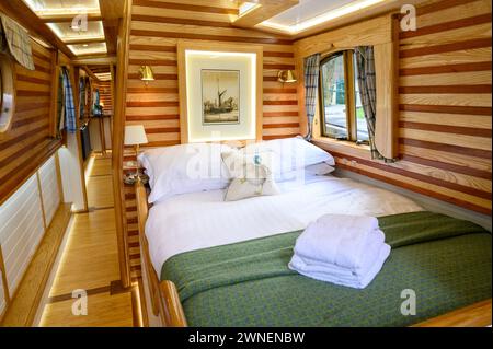 Komfortabel aussehendes Doppelbett in einem Mietboot auf den Binnenwasserstraßen in Wales mit Bettwäsche und einem Haufen Handtücher auf dem Bett. Stockfoto