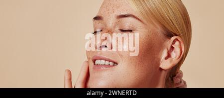 Seitenansicht einer lächelnden blonden Frau mit Sommersprossen, die ihr Gesicht berühren. Atelier-Nahaufnahme einer jungen positiven Frau mit geschlossenen Augen gegen Pastell Backgr Stockfoto