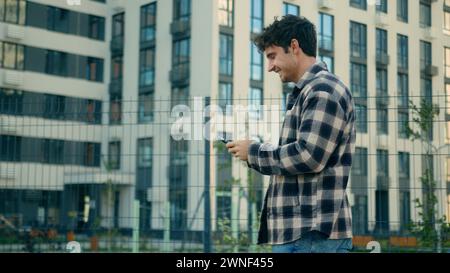 Seitenansicht glücklicher kaukasischer Mann lässige Fußgängerstraße in der Nähe moderner Gebäude in der Stadt männlicher Geschäftsmann lächelnder Kerl, der auf Handy-Smartphone schaut Stockfoto