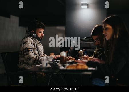 Drei konzentrierte Freiberufler arbeiten nachts in einem schwach beleuchteten Café an ihren Laptops, umgeben von Kaffee und Gebäck. Stockfoto