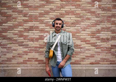 Porträt eines stilvollen Mannes, der mit kabellosen Kopfhörern glücklich in die Kamera schaut, lehnt sich gegen eine Ziegelwand. Stockfoto