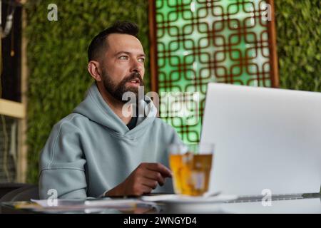 Nachdenklicher bärtiger Mann in legerer Kleidung im Café mit Laptop Stockfoto