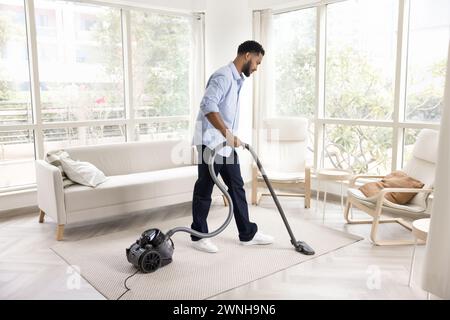 Ernster junger afrikanischer Hausbesitzer saugt Boden im Wohnzimmer Stockfoto