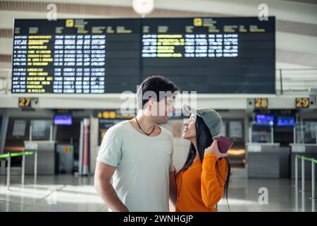 Junge Paare von Reisenden zeigen ihren Reisepass vor den Informationsbildschirmen des Terminals und schauen sich verliebt an Stockfoto