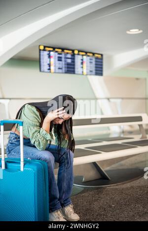 Vertikales Portrait verzweifeltes und gestresstes lateinmädchen am Flughafen, das auf der Bank sitzt und ihre Koffer weint Stockfoto