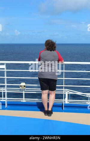 Eine Frau mittleren Alters auf einem Kreuzfahrtschiff, die sich an ein Geländer lehnt und auf das Meer blickt Stockfoto