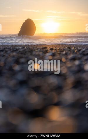 Ein felsiger Strand mit einem großen Felsen im Vordergrund und einem Sonnenuntergang im Hintergrund. Die Sonne untergeht hinter dem Felsen und strahlt ein warmes Leuchten über den Stockfoto