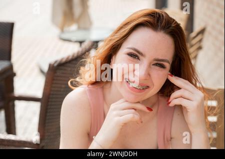 Schöne junge rothaarige Frau mit Zahnspangen an den Zähnen lächelnd, während sie in einem Café im Freien sitzt. Stockfoto