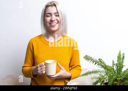 Eine Frau in einem leuchtend gelben Pullover, die Augen mit einem zufriedenen Lächeln geschlossen, genießt einen Moment mit ihrer Kaffeetasse und weckt ein Gefühl von Wärme und Glück. Stockfoto