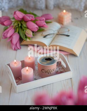 Eine Tasse Kaffee mit einem Buch und Gläsern, Tulpen, Kerzen im Innenraum, ein ästhetisches Foto Stockfoto