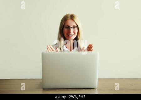 Glückliche junge Frau, die an Laptop-Begrüßungen arbeitet, kommuniziert mit Freunden per Videoanruf, die am Schreibtisch im Büro oder zu Hause sitzt Stockfoto