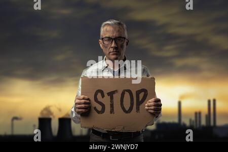 Schmutziger, verzweifelter Geschäftsmann, der ein Schild hält und gegen Verschmutzung, Industrieanlagen und giftige Gase im Hintergrund protestiert Stockfoto