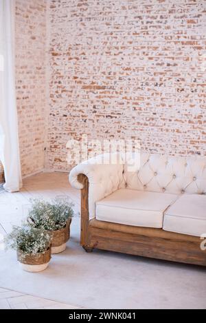 Inneneinrichtung mit einem weißen gesteppten Sofa mit Holzsockel vor einer leeren Ziegelwand, mit weißen Vorhängen und Blumentöpfen auf einem grauen Teppich Stockfoto