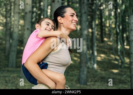 Mutter und Tochter teilen sich eine fröhliche Umarmung während einer Huckepack-Fahrt in einem sonnendurchfluteten Wald und genießen ihre gemeinsame Familienzeit - Familienurlaub pur Stockfoto