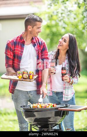 März. 02. 2024 Liptovsky Mikulas. Slowakei - Frau bringt ihrem Mann eine Flasche Cola, während er auf dem Grill im Garten grillt. Stockfoto