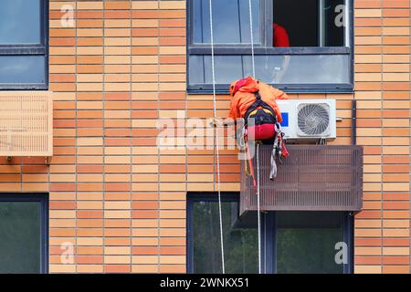 Ein funktionierender Kletterer in Uniform verbindet die Klimaanlage unter dem Fenster eines Wohnhauses Stockfoto