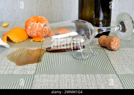 Das Glas liegt in einer Weinpfütze auf dem Tisch, daneben stehen eine leere Flasche und Obst. Stockfoto