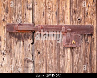 Eine alte Holztür mit einem aufklappbaren alten Eisenriegel. Rostiges massives Eisenschloss an einem verlassenen Holztor. Sonnenverblichenes, gereiftes und von Rinde gegessen Holz Stockfoto