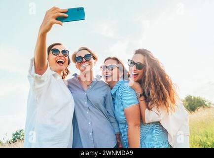Porträt von vier fröhlichen, lächelnden Frauen in einer Sonnenbrille, die sich zusammenschließen und Selfie-Fotos mit einem modernen Smartphone machen, während sie im Freien spazieren gehen. Die der Frau Stockfoto