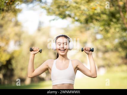 Eine temperamentvolle junge Frau genießt ihr Training in einem sonnigen Park und hebt Hanteln mit Kopfhörern Stockfoto